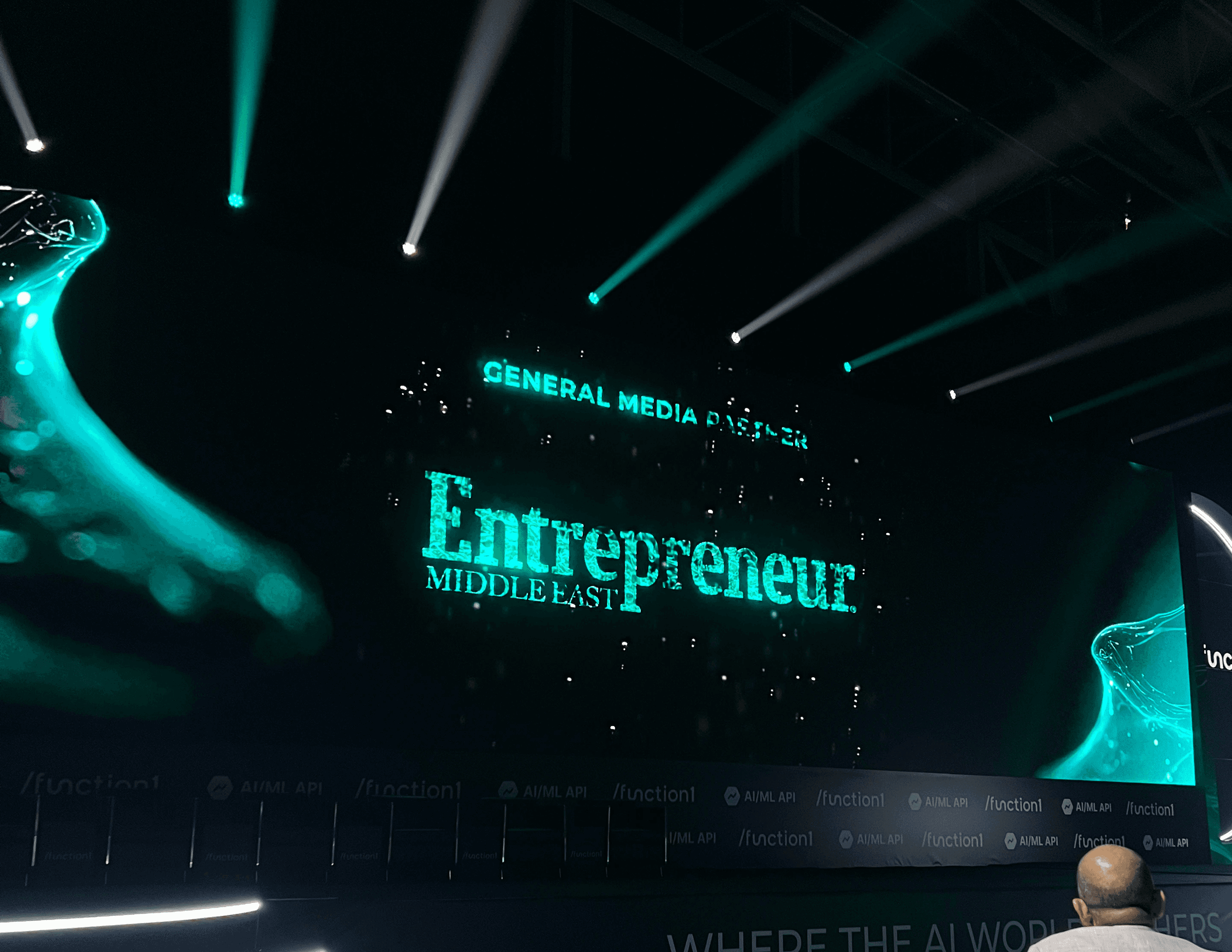 A dynamic stage with bright spotlights showcases a large display screen featuring the words "General Media Partner Entrepreneur Middle East" in bold text, set against a futuristic backdrop illuminated with green and white digital patterns.