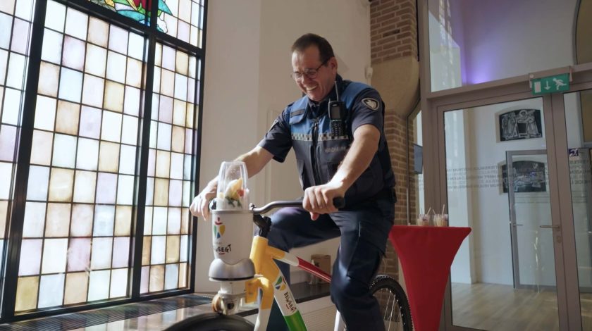 A person in a blue uniform is cycling on a stationary bike indoors, beside large stained-glass windows, near a red cocktail table with a drink on it.