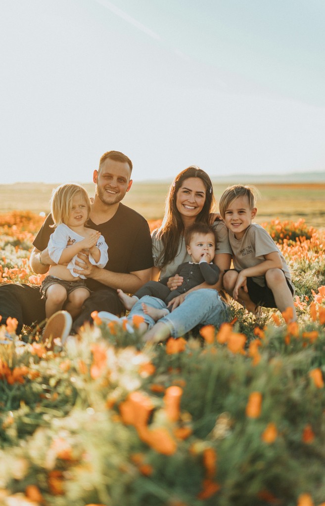 A cheerful family sitting together in a field of vibrant orange flowers, enjoying a sunny day, with the parents holding their three young children and smiling at the camera.