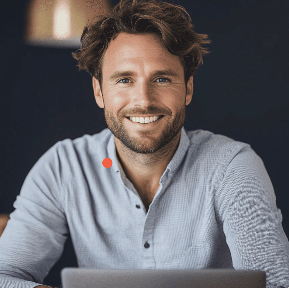 A smiling man with curly hair is sitting at a desk, looking at the camera, with a laptop and a lamp in the background.