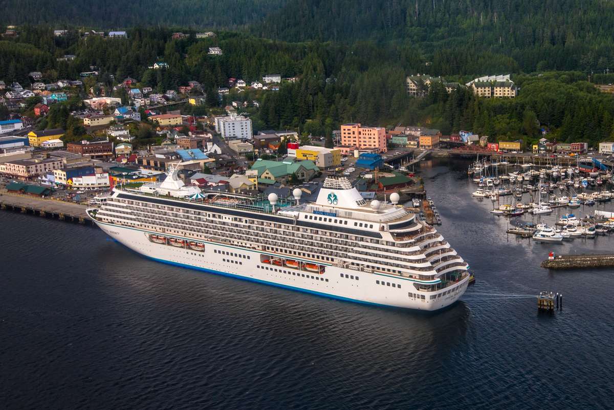 A large white cruise ship with a green and yellow funnel sails past a coastal town with colorful buildings and trees.