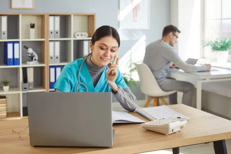 Decision support software used by a nurse managing medication records and calls with a laptop and paperwork.