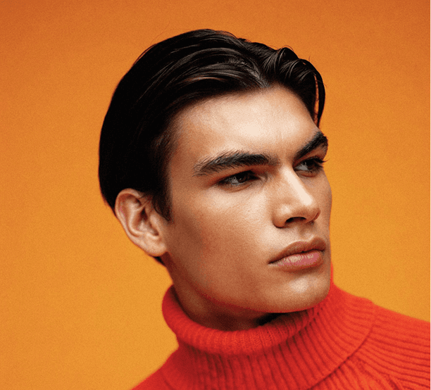 A man dressed in an orange sweater layered over a black turtleneck, posing casually.