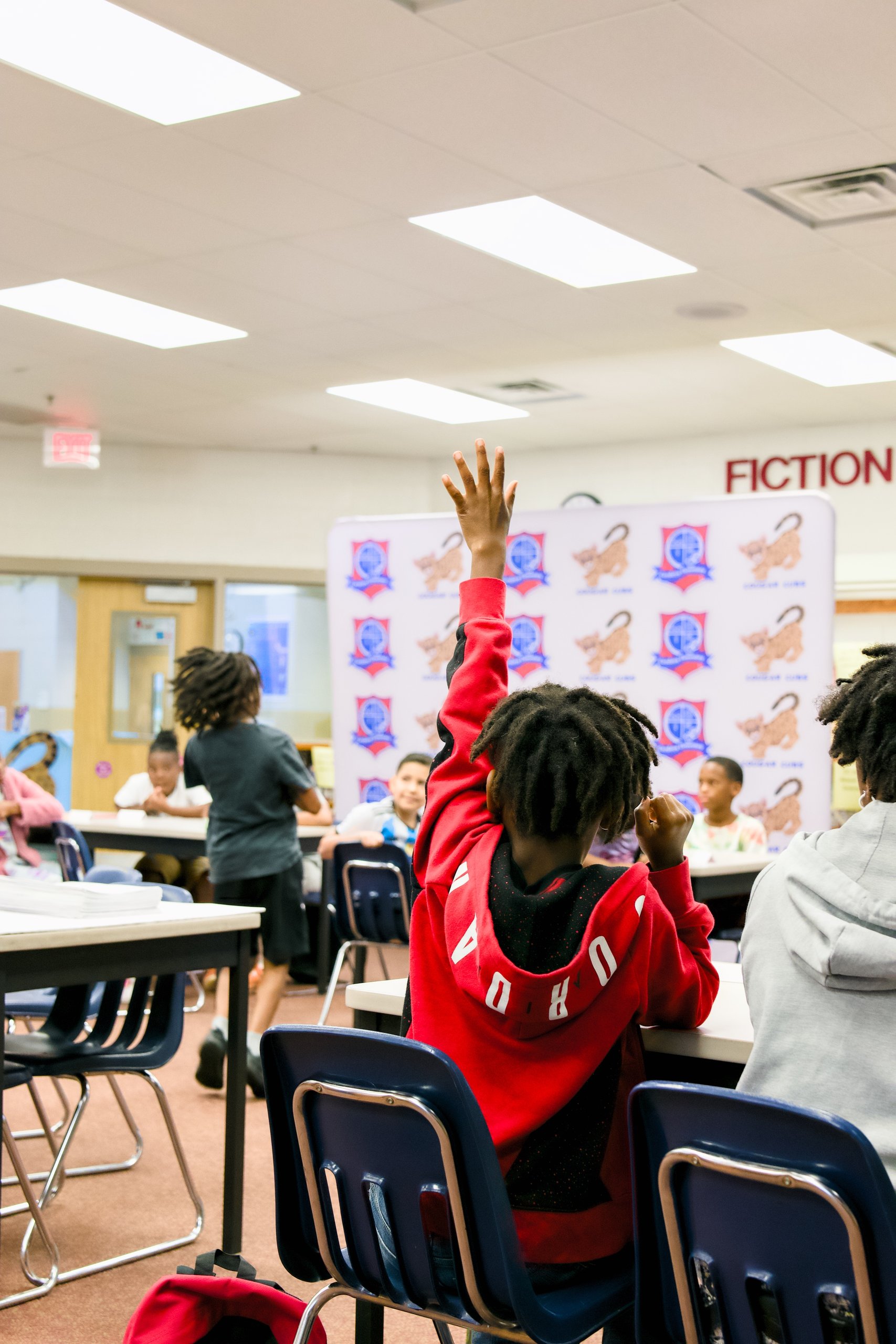 Child in a red hoodie raises his hand in a school library