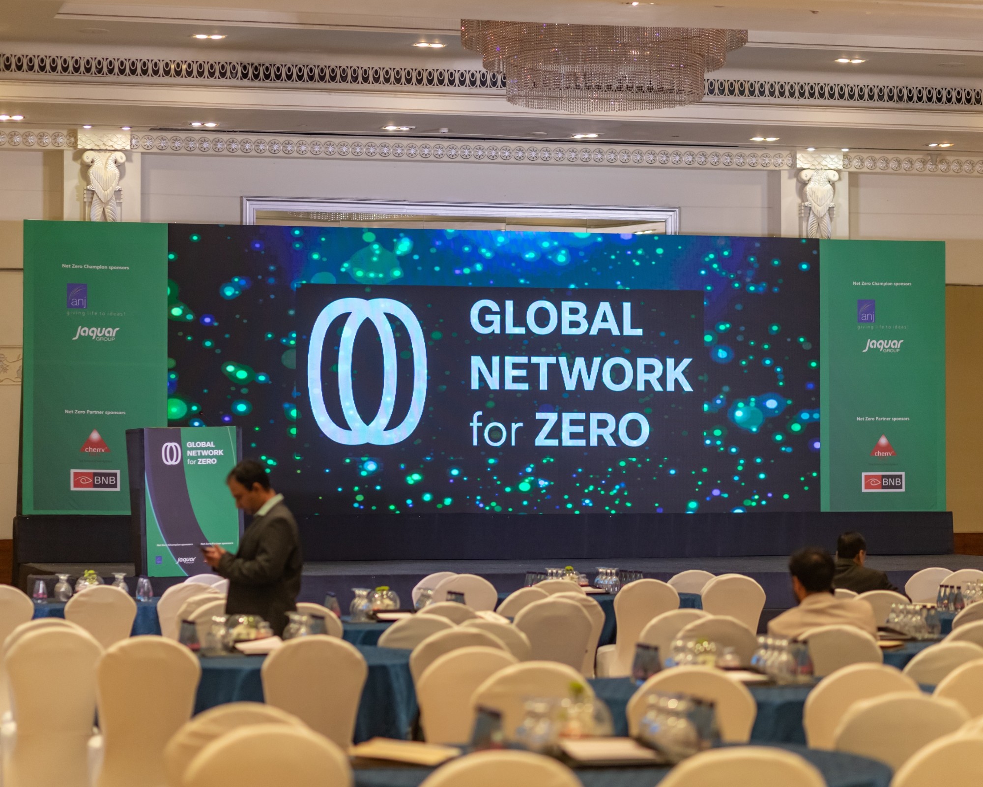 A man stands while two people sit at a table in an office setting, smiling and engaged in conversation. The mood is friendly and collaborative.