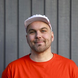 Kasvokuva Värisalmen yrittäjästä, hymyilevä ilme ja katse kameraan, päällä oranssi työpaita ja maalarinlakki, pystylaudoituksen edessä.