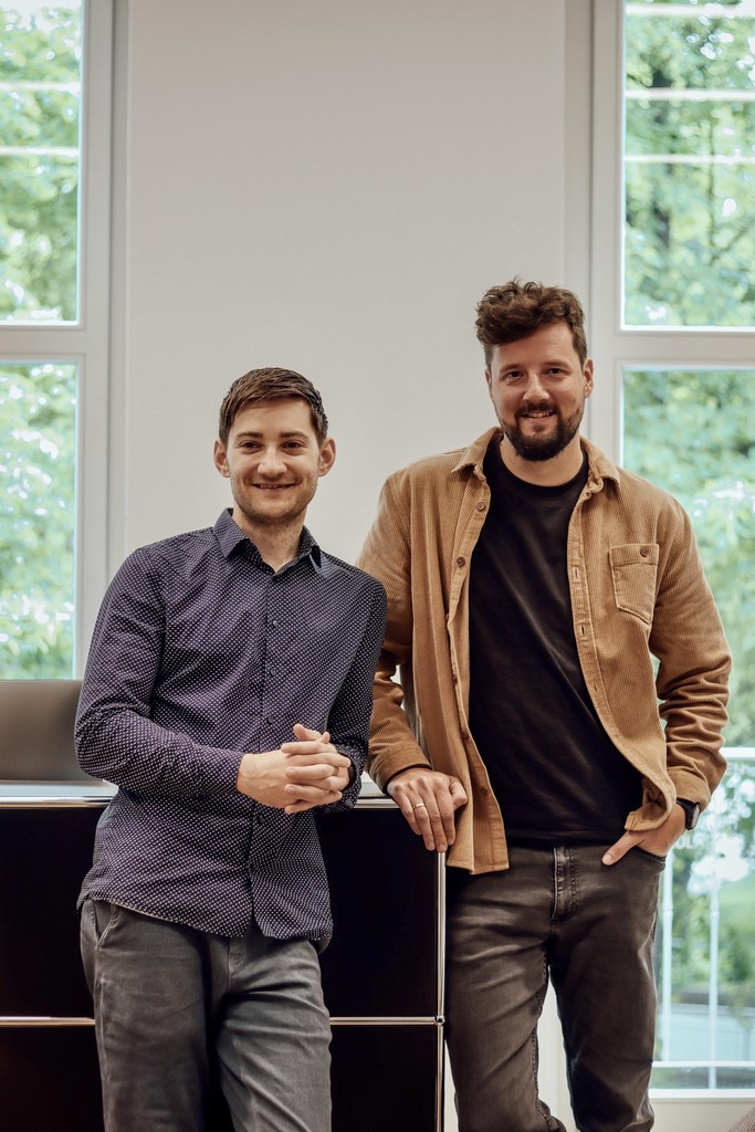 Two men stand together, smiling and shaking hands, with a bright window in the background.