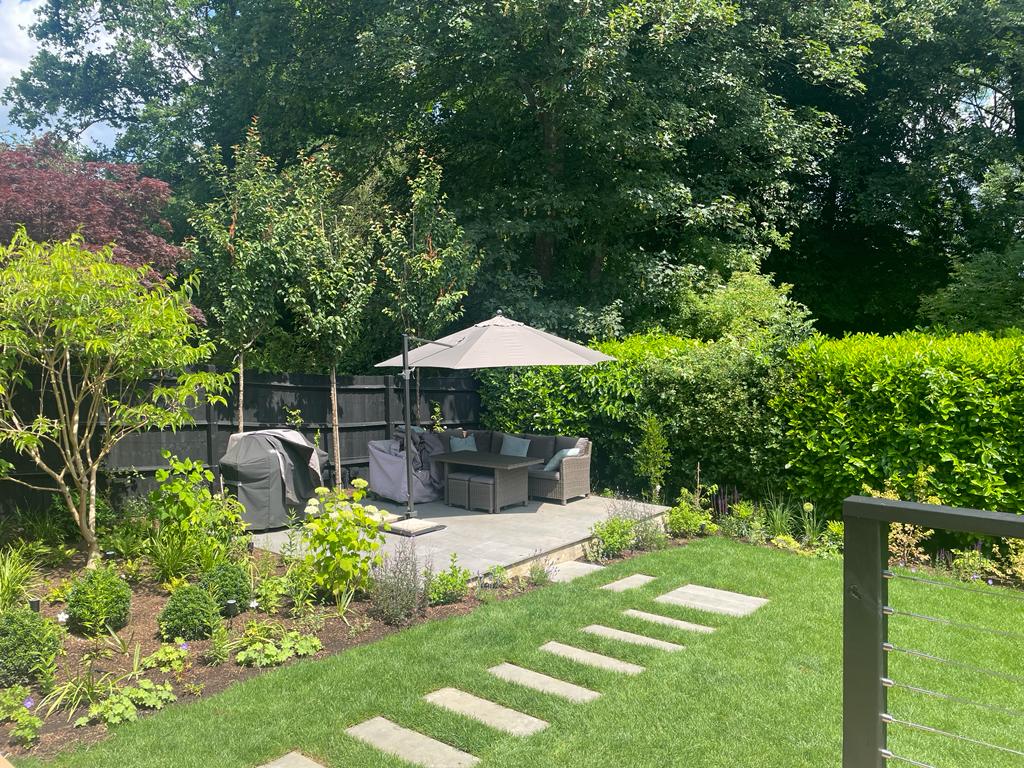 Lush garden scene featuring a gazebo under a parasol, surrounded by greenery and flower beds.