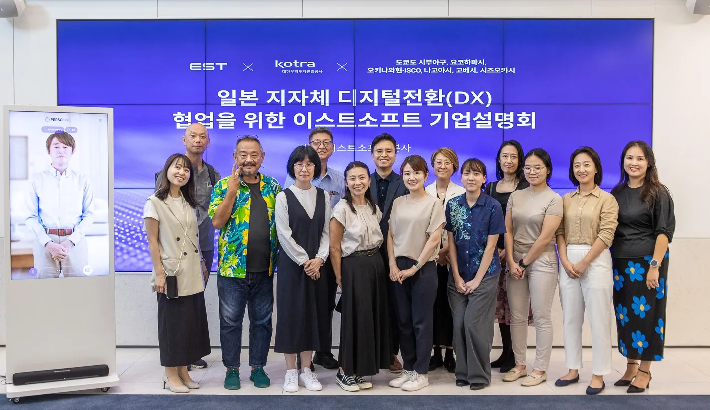 ภาพถ่ายหมู่ของเจ้าหน้าที่เทศบาลญี่ปุ่นและตัวแทนของ ESTsoft ด้านหน้าจอการนำเสนอและตู้ AI Human ที่สำนักงานใหญ่ของ ESTsoft