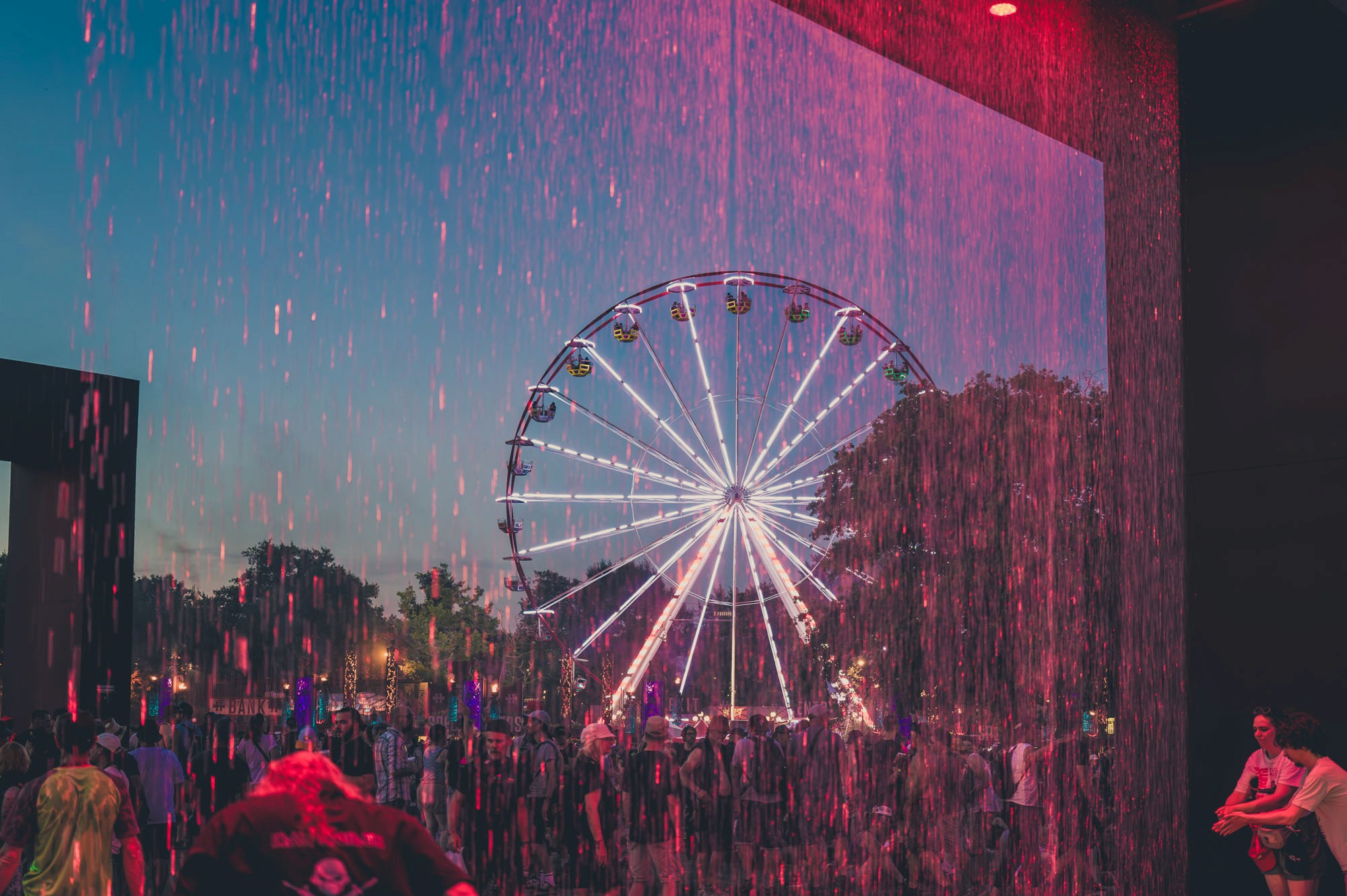 Grande roue illuminée vue à travers un rideau d'eau teinté de rose, tombée de la nuit