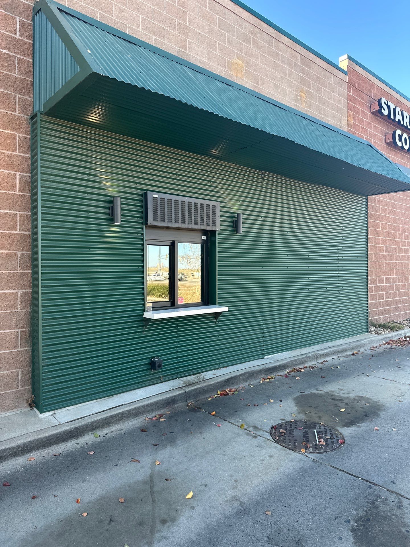 Drive-thru window area at a Starbucks store featuring corrugated green metal siding, overhead awning, and clean pavement.