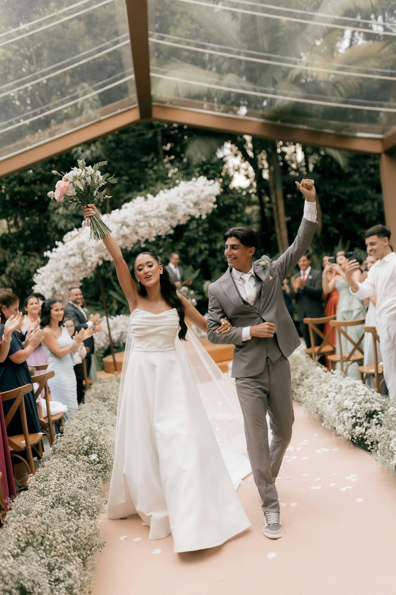 Newlyweds walking triumphantly down a flower-lined aisle under a glass canopy with cheering guests and white orchids behind them