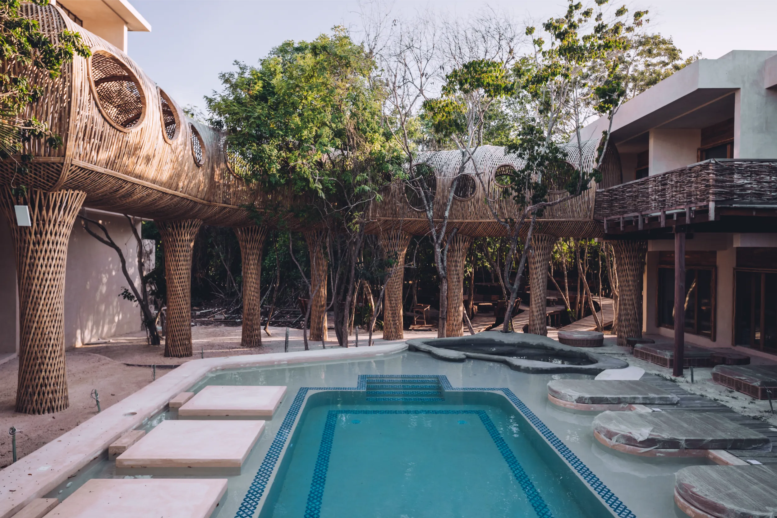 Wide view of the Cervecería Tulum pool area, featuring the elevated bamboo tunnel supported by organic woven columns that frame the recreational space.