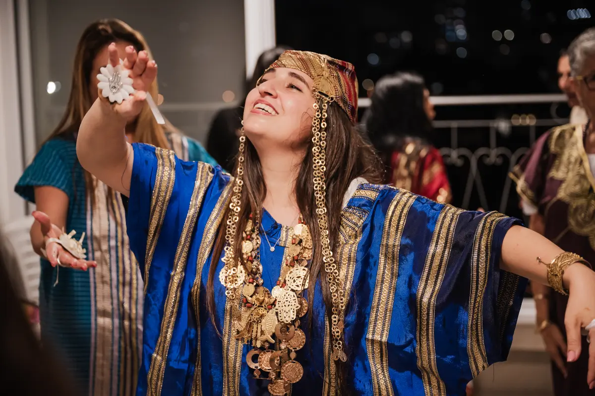 צילום תקריב מרגש מתוך טקס חינה מסורתי. אישה צעירה בלבוש כפתן כחול מעוטר בזהב ובתכשיטים מסורתיים רוקדת בעיניים עצומות ובחיוך של אושר, מאת Shiriz Photography והצלמת שירי עציץ