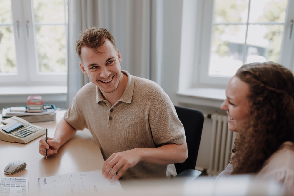 Zwei Personen sitzen nebeneinander am Tisch und gehen gemeinsam Dokumente durch.