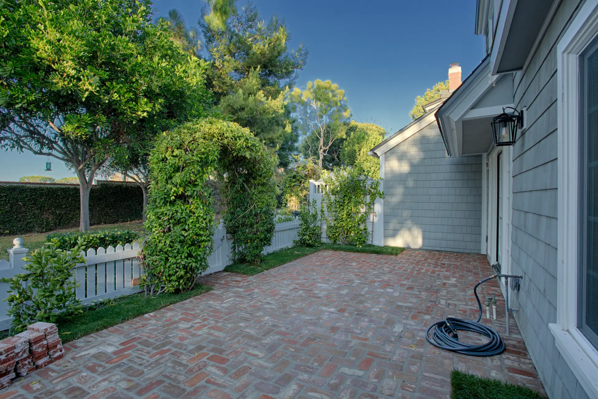 Side patio of a Cape Cod style home, with a brick patterned path and lush greenery along the white fence. From Dana Point Cape Cod remodel. Photo by Chris Darnall.
