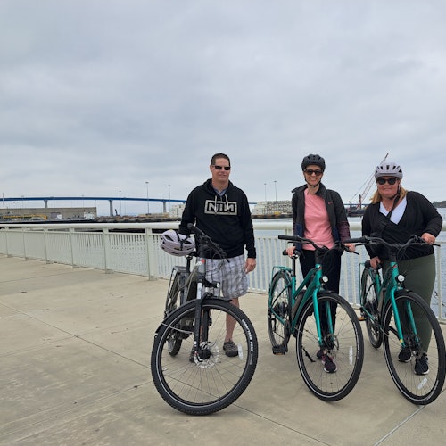 三个人在滨水走道上骑自行车。 他们戴着头盔，穿着休闲服装。 背景中有一座桥。