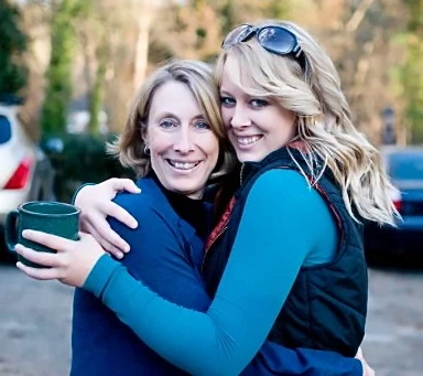 Two women embrace outside, one holding a green mug.