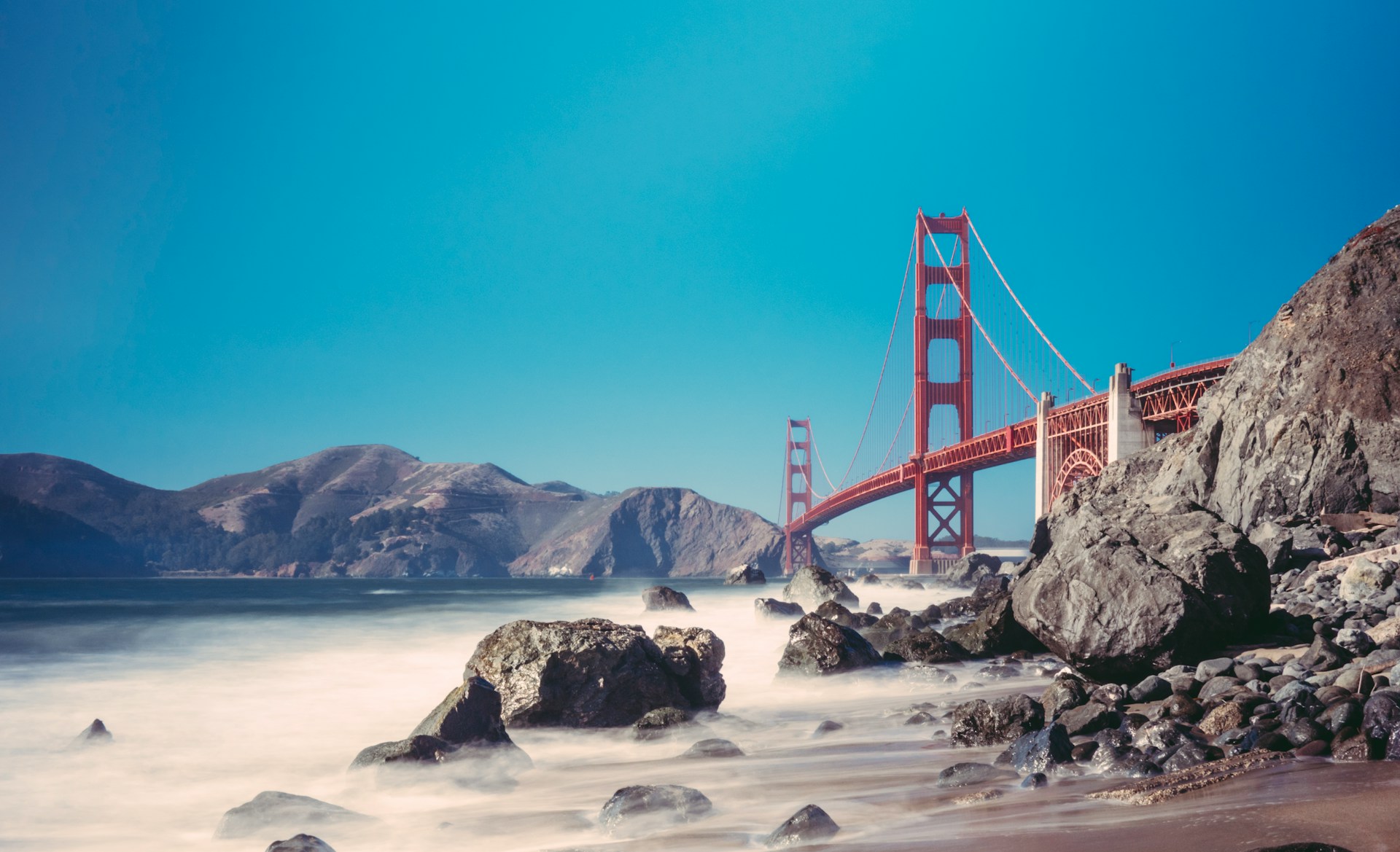 Golden Gate Bridge viewed from a rocky beach with long-exposure smooth water.