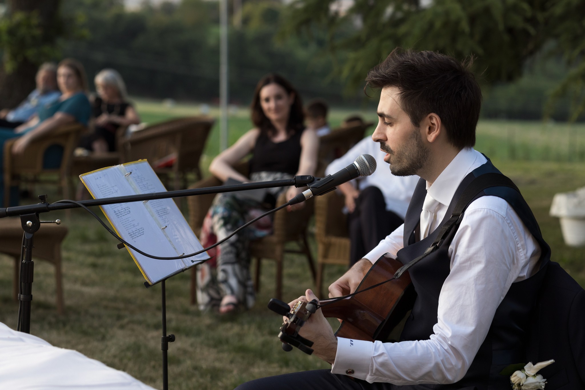 Matteo canta con chitarra al matrimonio di Francesca a Villa Peroni Riva, ospiti seduti sullo sfondo nel giardino
