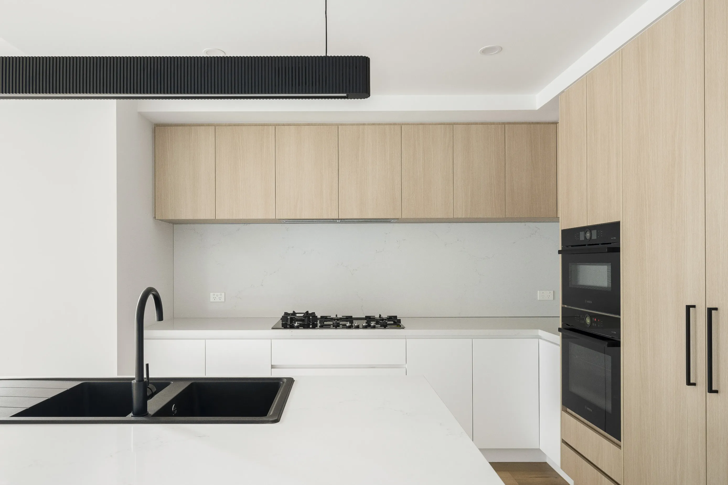 Minimalist kitchen with pale-oak overhead cabinetry, white base cabinets, a marble-look splashback, gas cooktop, black wall ovens, a black fluted linear pendant and a white island with black undermount sink and tap
