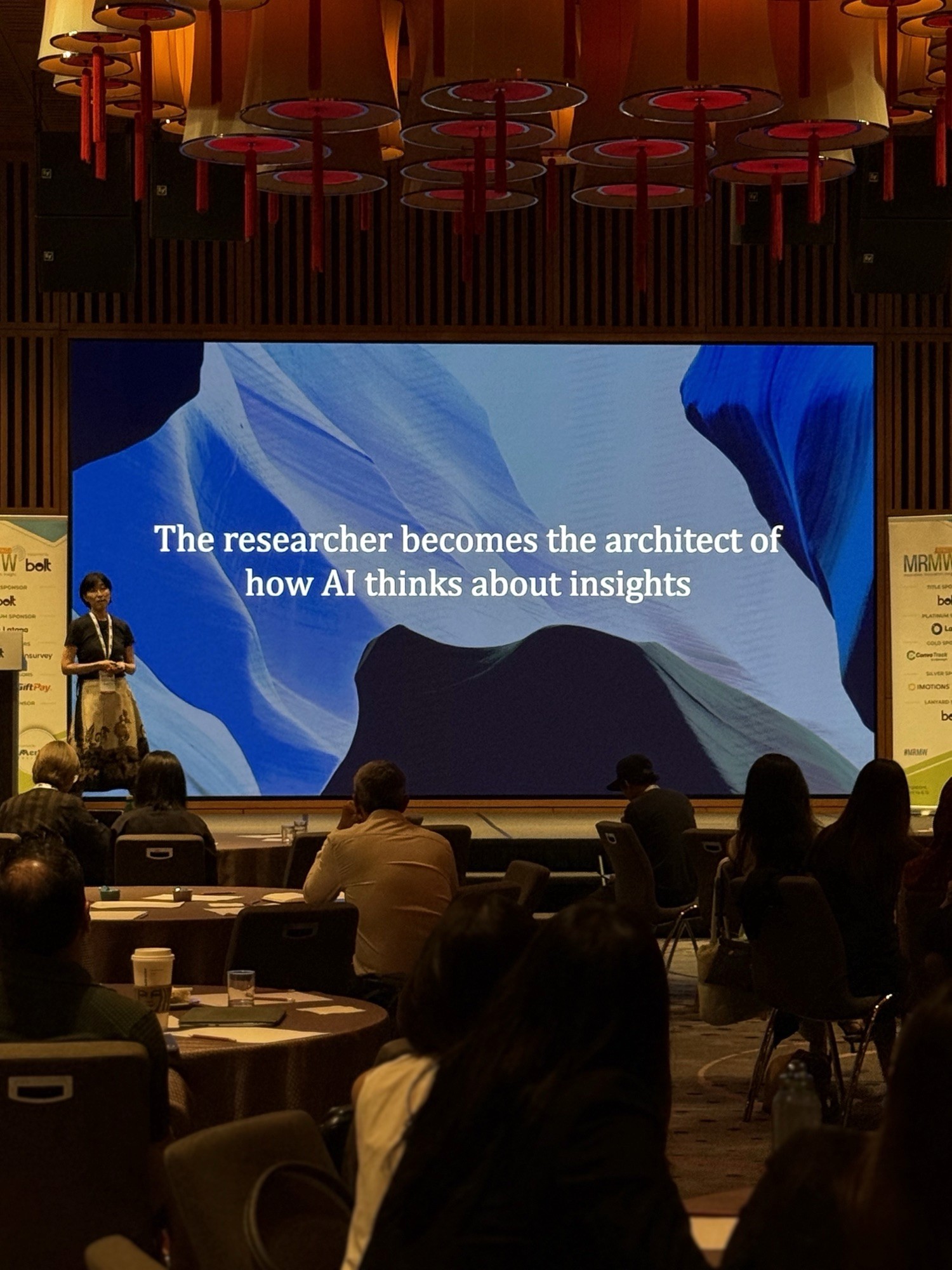 A large screen displays a blue background with text, while attendees sit in a conference setting.