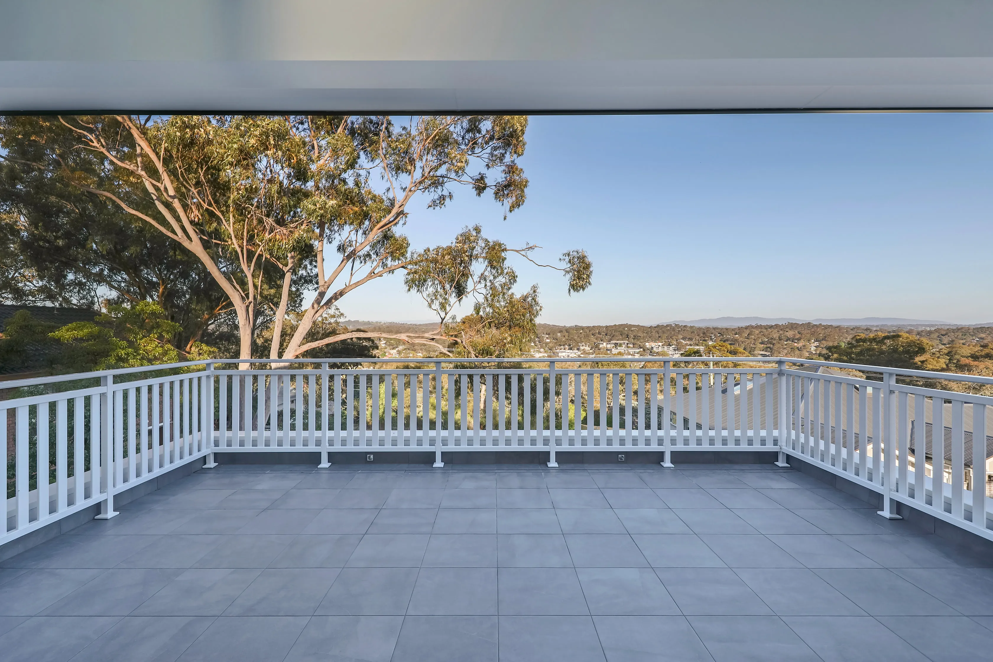 Tiled upper-level balcony with a white timber balustrade overlooking gum trees and a leafy valley under clear sky