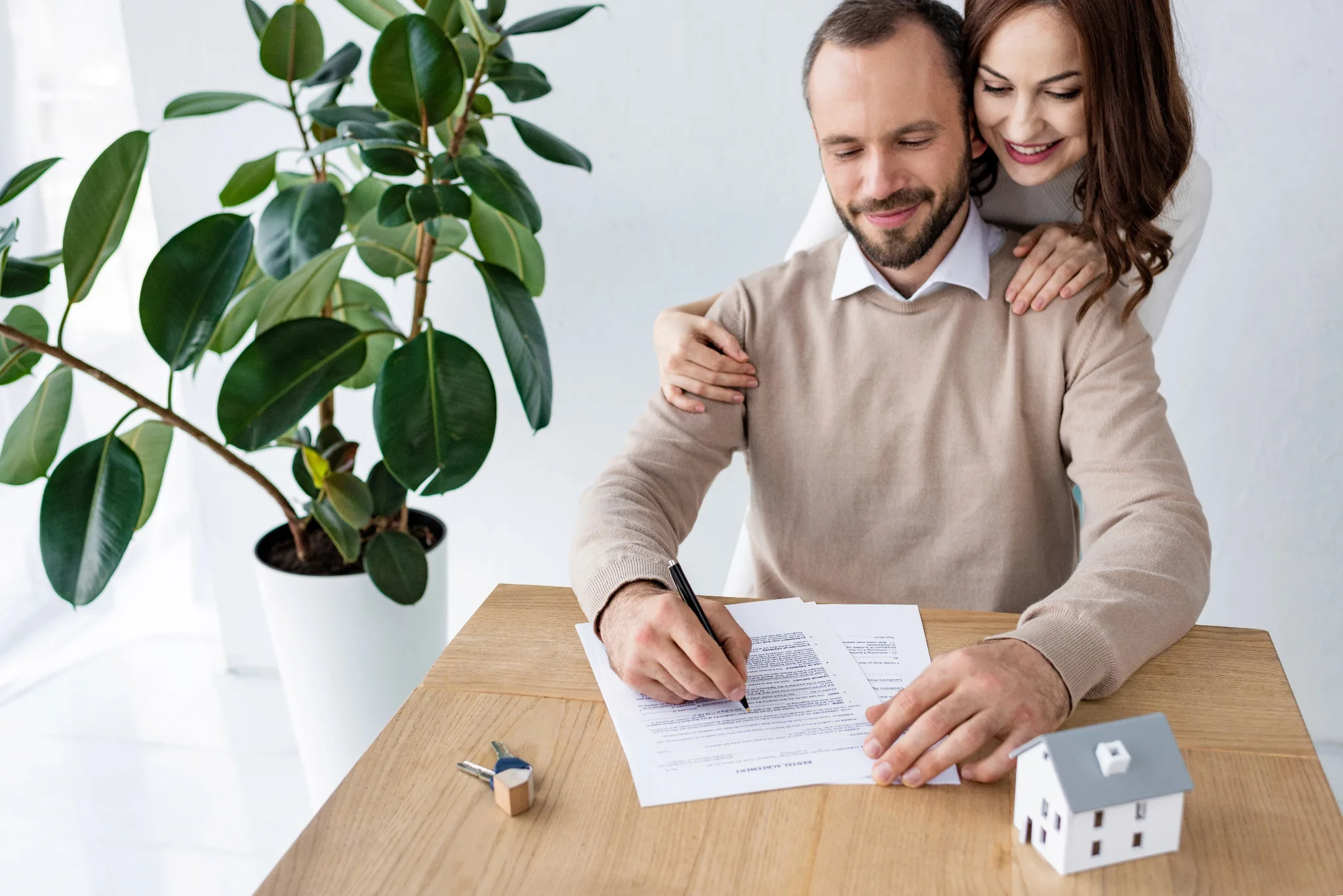 A couple signs a real estate document at a wooden table with a small house model and keys, with a potted plant visible in the background.