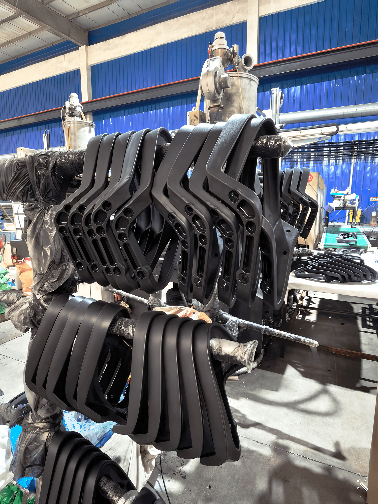 A close-up view of the office chair armrest production line at SingOFai Furniture. Several rows of matte black, molded plastic armrests are hung neatly on horizontal metal racks for drying or storage. The armrests feature a modern, angular ergonomic design. In the background, the industrial factory setting includes blue corrugated metal walls, various manufacturing machinery, and additional components on a nearby worktable.