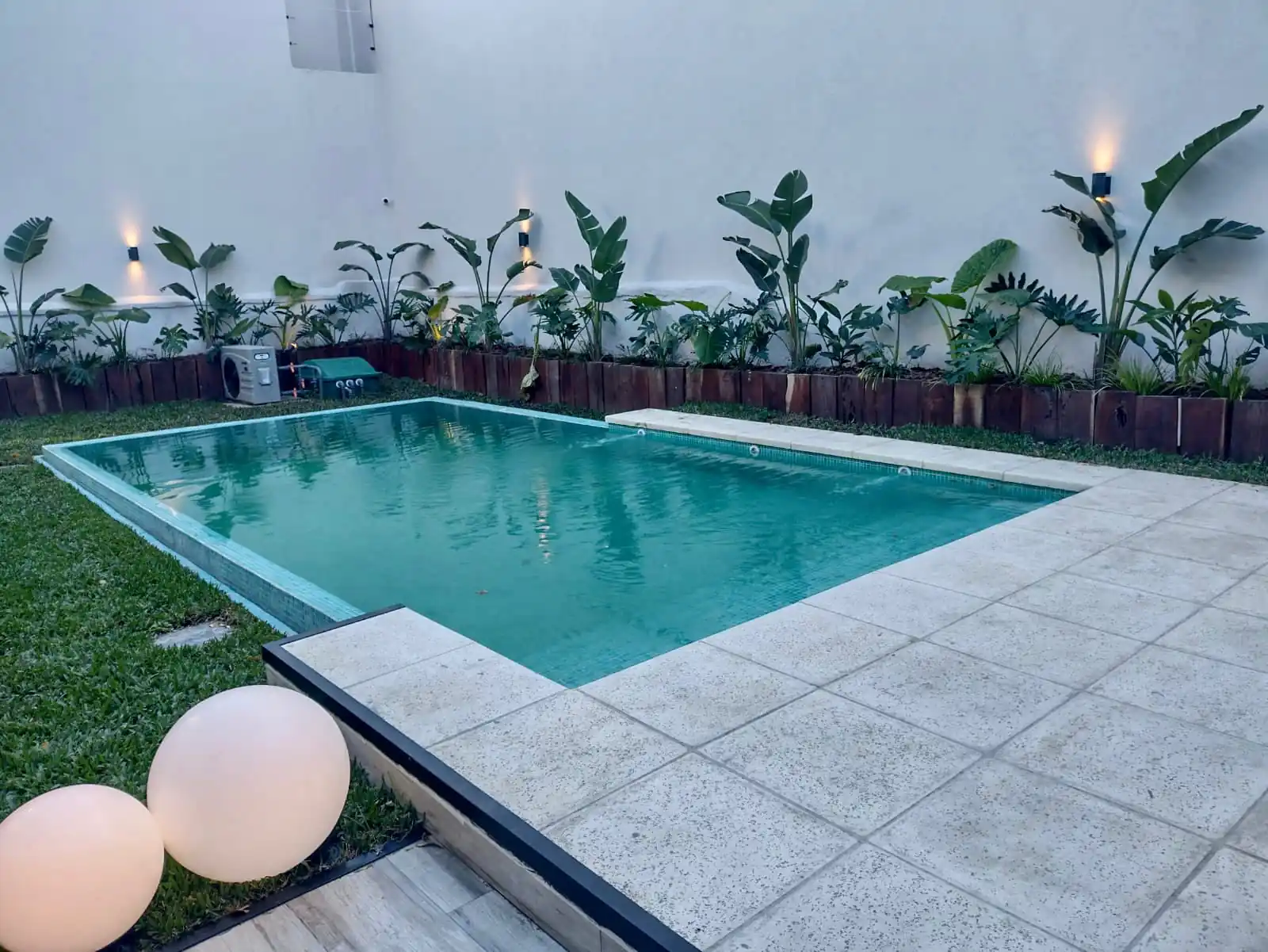 Moderna piscina de patio rodeada de baldosas claras, destacando esferas luminosas decorativas y un cantero con plantas tropicales iluminadas.