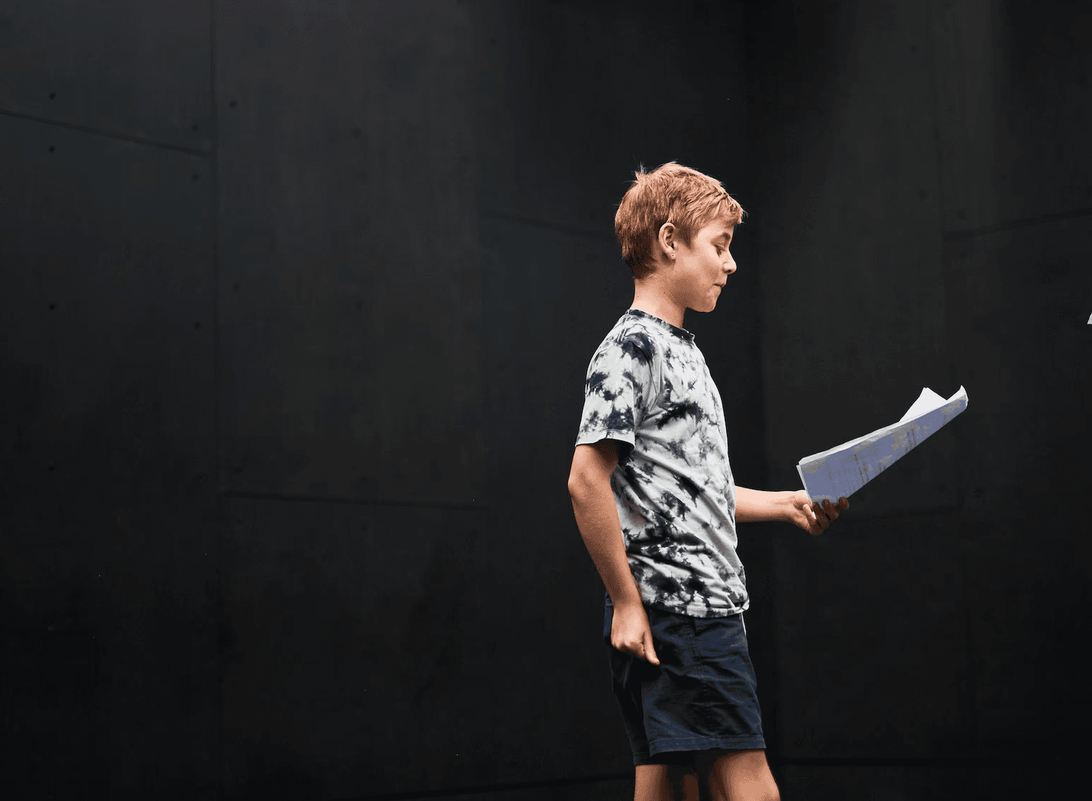 A child stands alone rehearsing with a script in hand in a dark-walled studio.