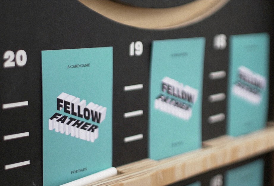 Four teal cards labeled “FELLOW FATHER” are displayed on a black board with numbered slots. The cards are arranged vertically and feature white text and graphics. A white stick is visible in the bottom left corner.