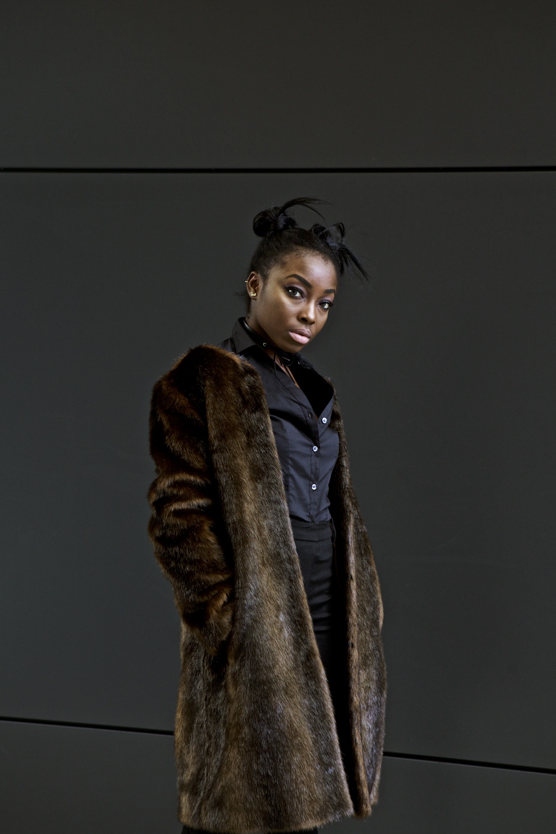 Studio portrait of a model in a dark outfit and oversized fur coat against a black background.