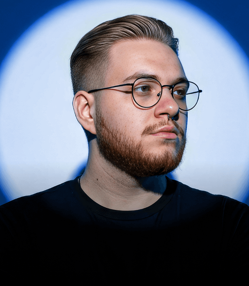 A young man with round glasses, a nose ring, and facial hair looks toward the camera against a dark, softly colored background.