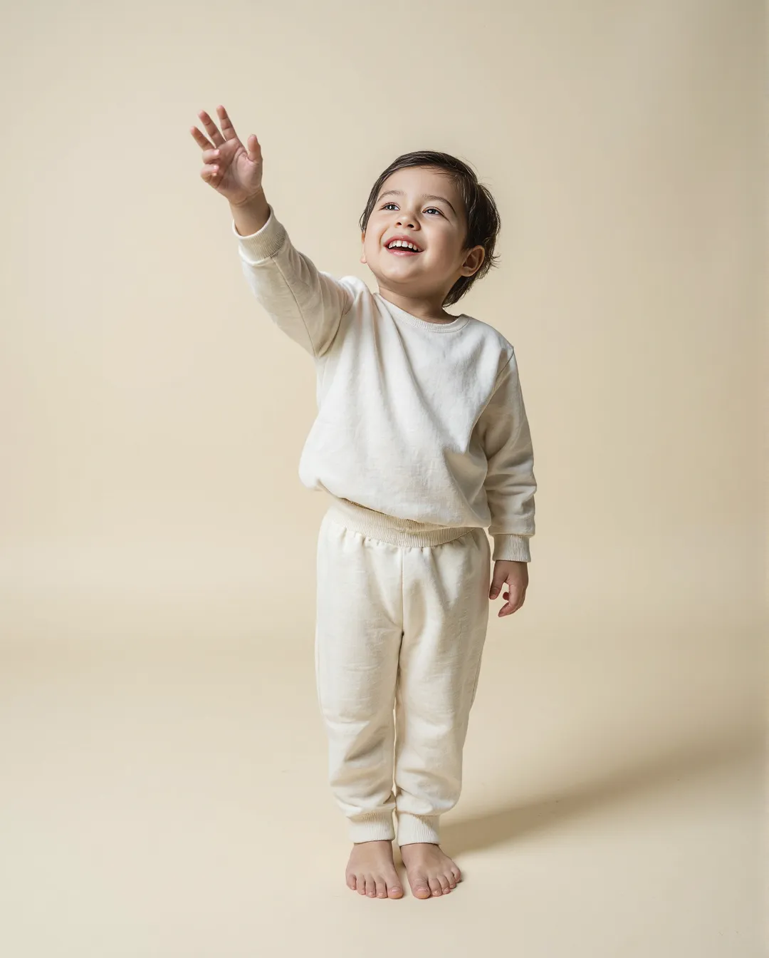 Child wearing cream loungewear set against neutral background, reaching upward with joyful expression, perfect for minimal catalogue styling