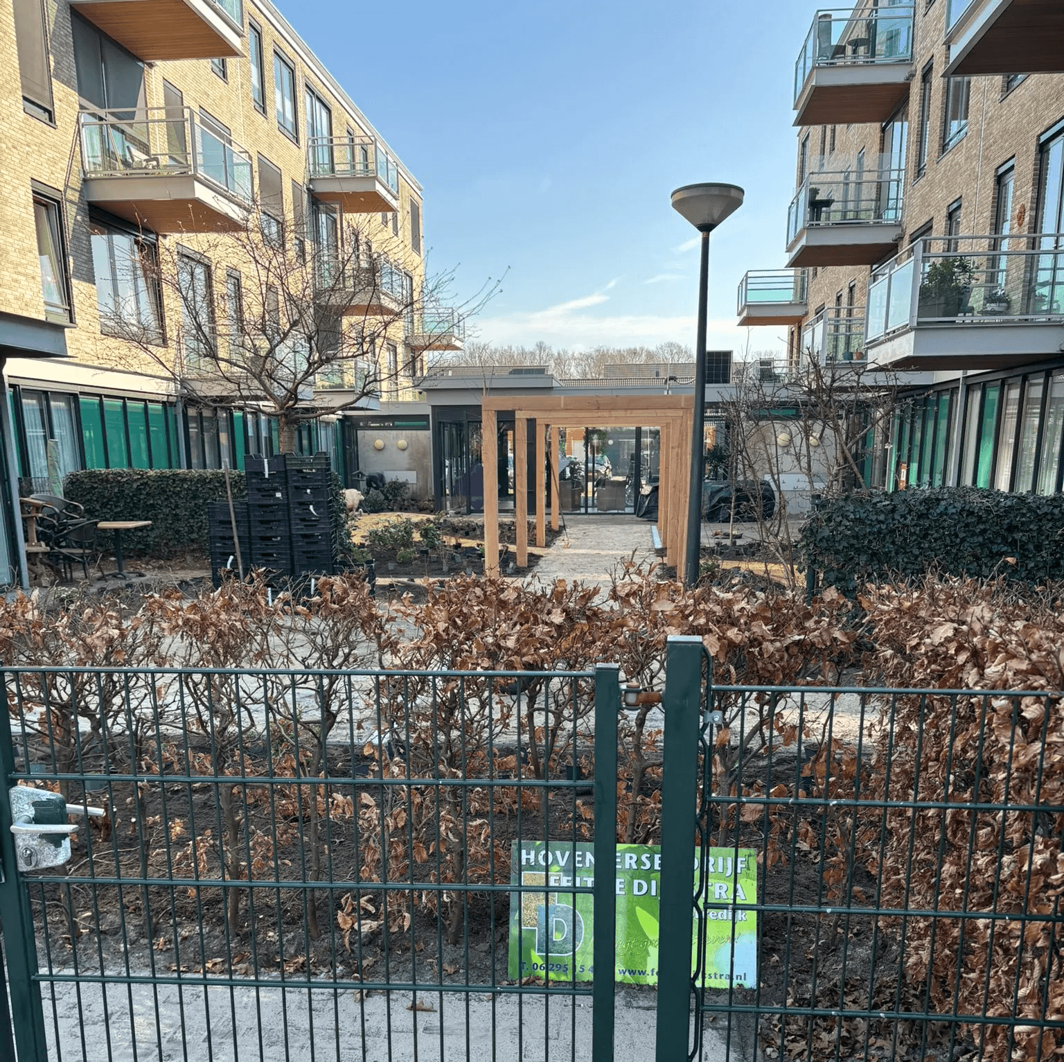 Belevingstuin met kale bomen, houten pergola, grindpad en groene hagen tussen zorgappartementen in Gorredijk.