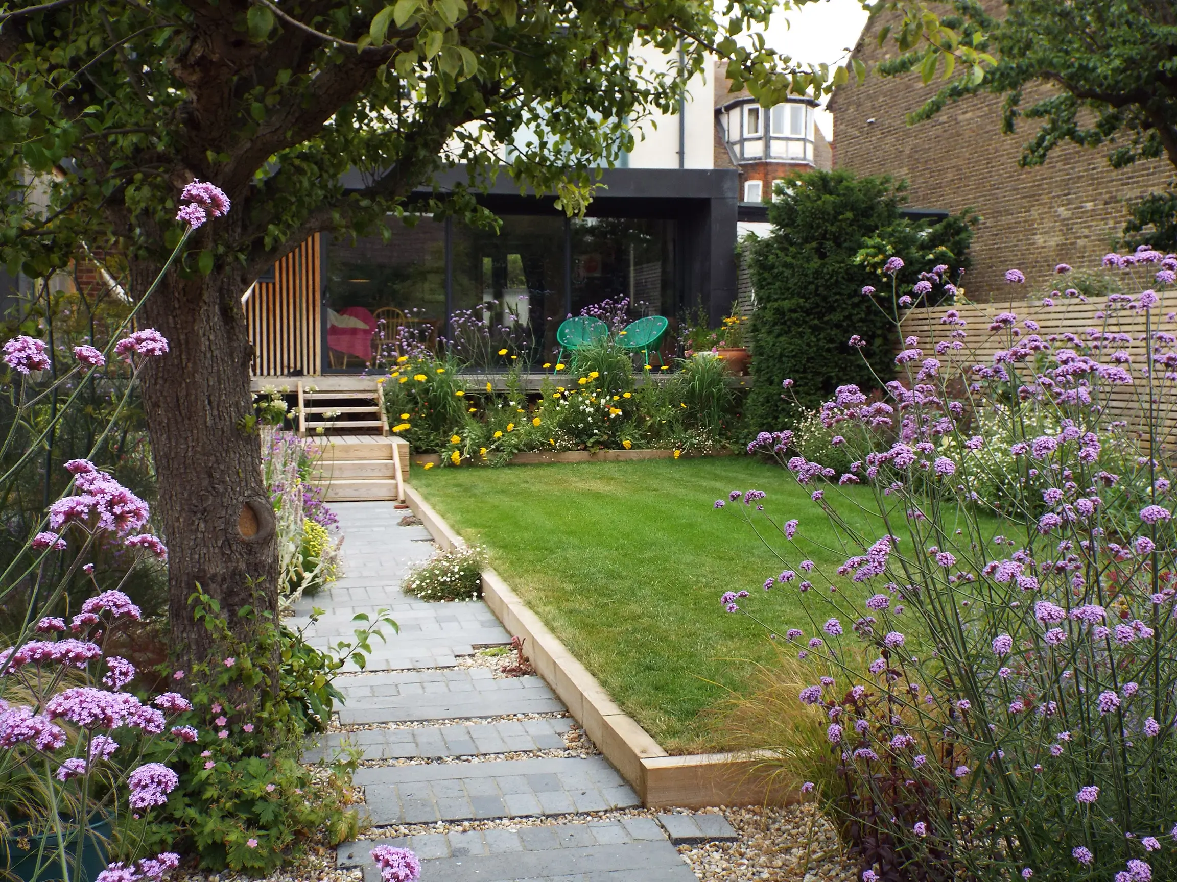 A serene garden pathway lined with flowers, leading to a grassy area and a glimpse of a home in the background.