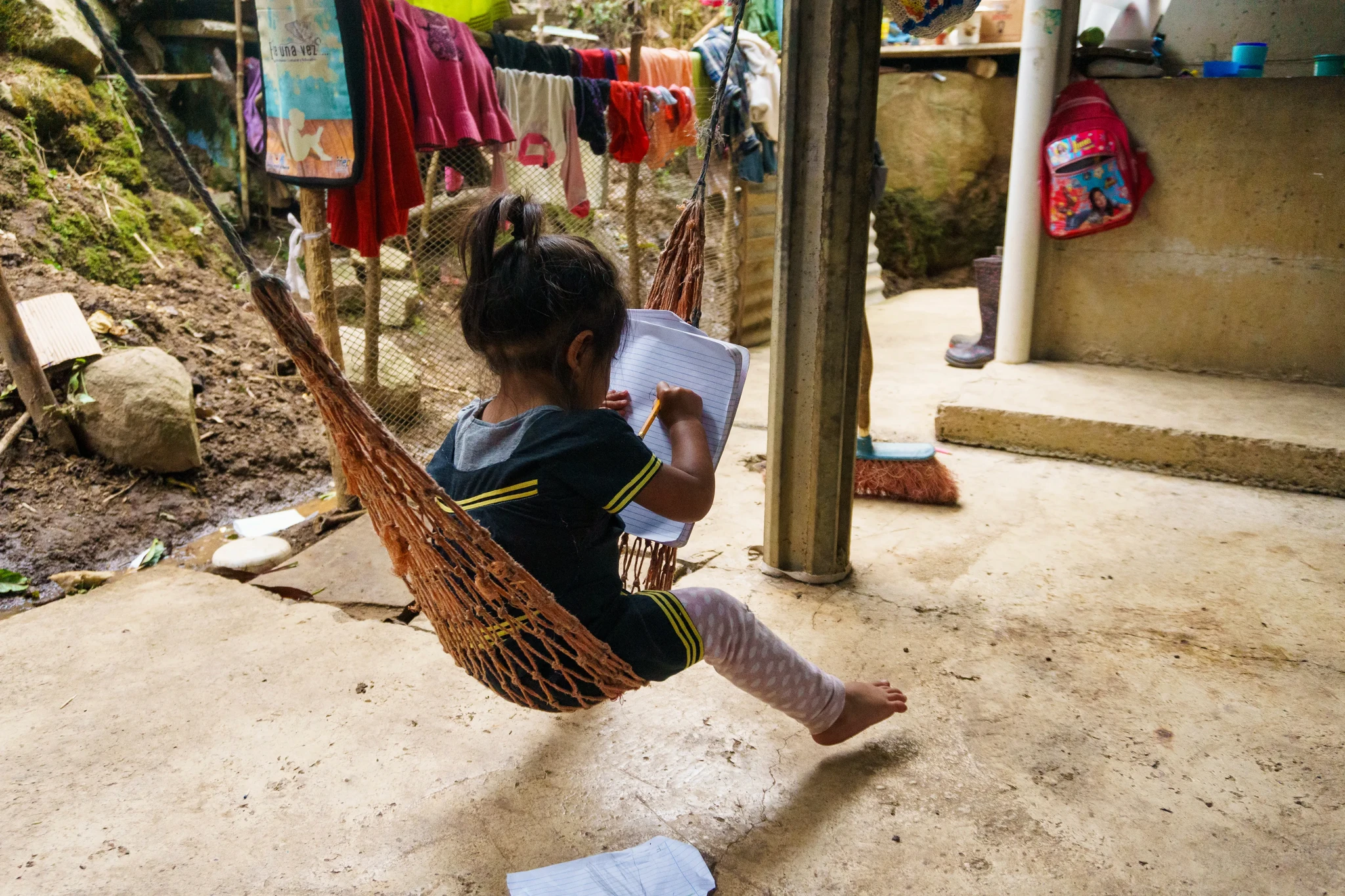 A young child sits in a hammock outdoors, writing in a notebook. The area has laundry hanging to dry and various household items are visible. The setting appears informal and rustic.