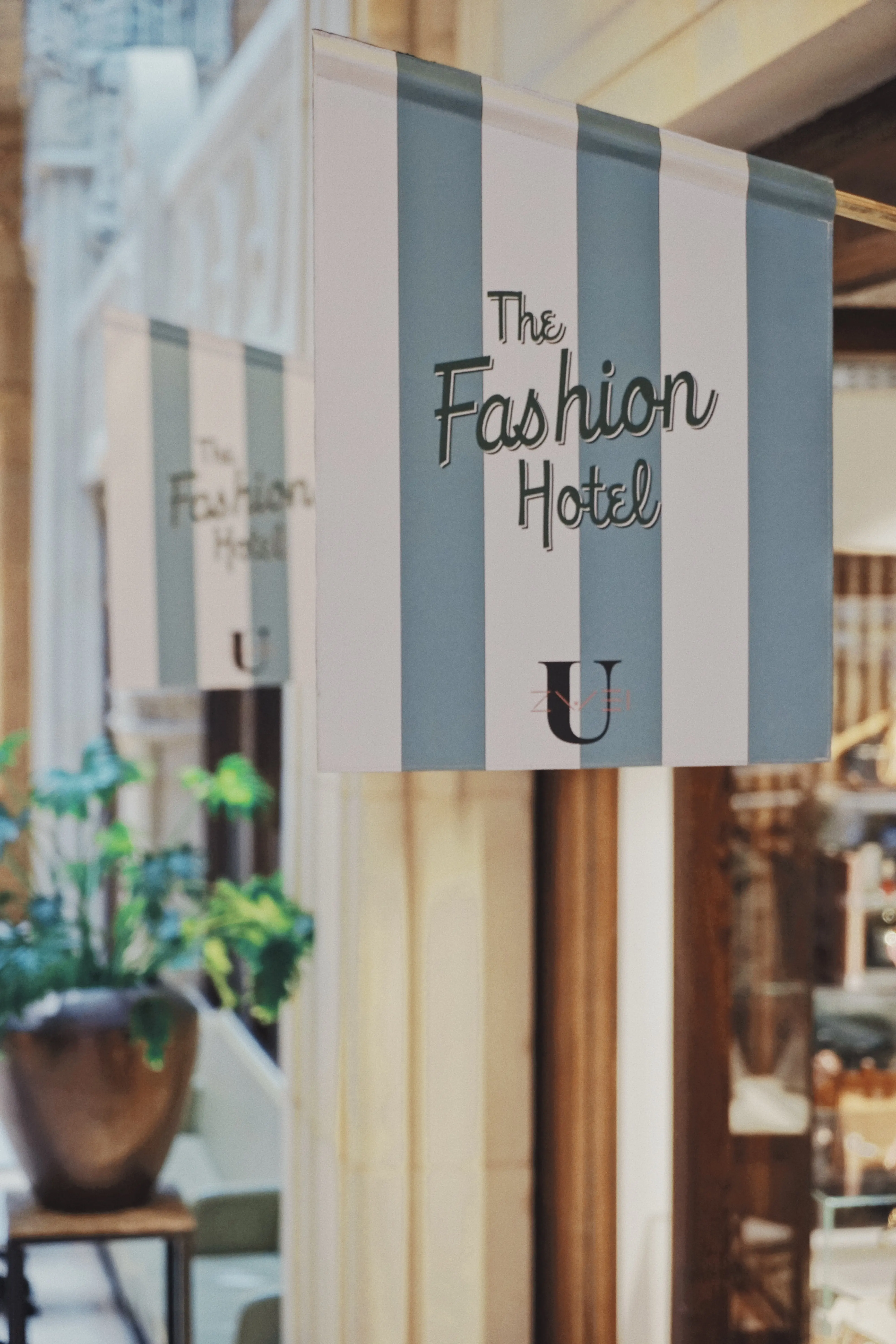 Striped blue-and-white flag signs reading The Fashion Hotel mounted on golden poles inside UZWEI, with plants and warm shop lighting in the background.