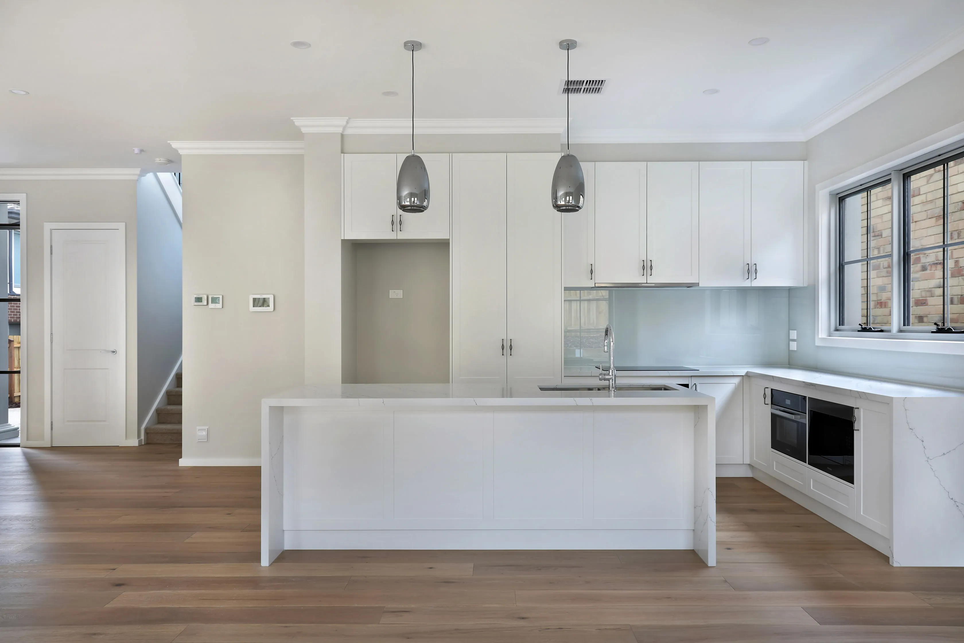 Open-plan kitchen with white shaker cabinetry, a Calacatta-look waterfall stone island, chrome dome pendants and oak floors, with a carpeted staircase beyond