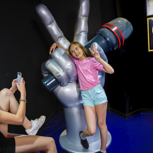 Dívka pózuje s gestem míru vedle obří sochy ruky držící mikrofon, zatímco ji jiná osoba fotografuje.