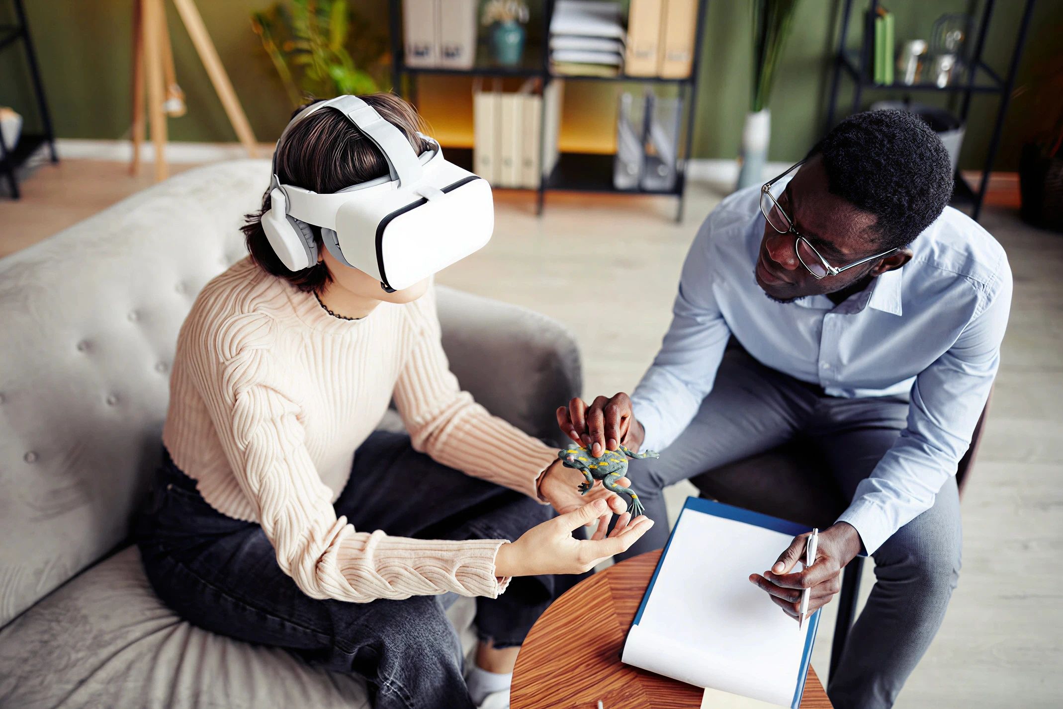 Patient wearing a VR headset during a VR therapy session for phobia treatment, holding a lizard toy while a therapist observes and takes notes