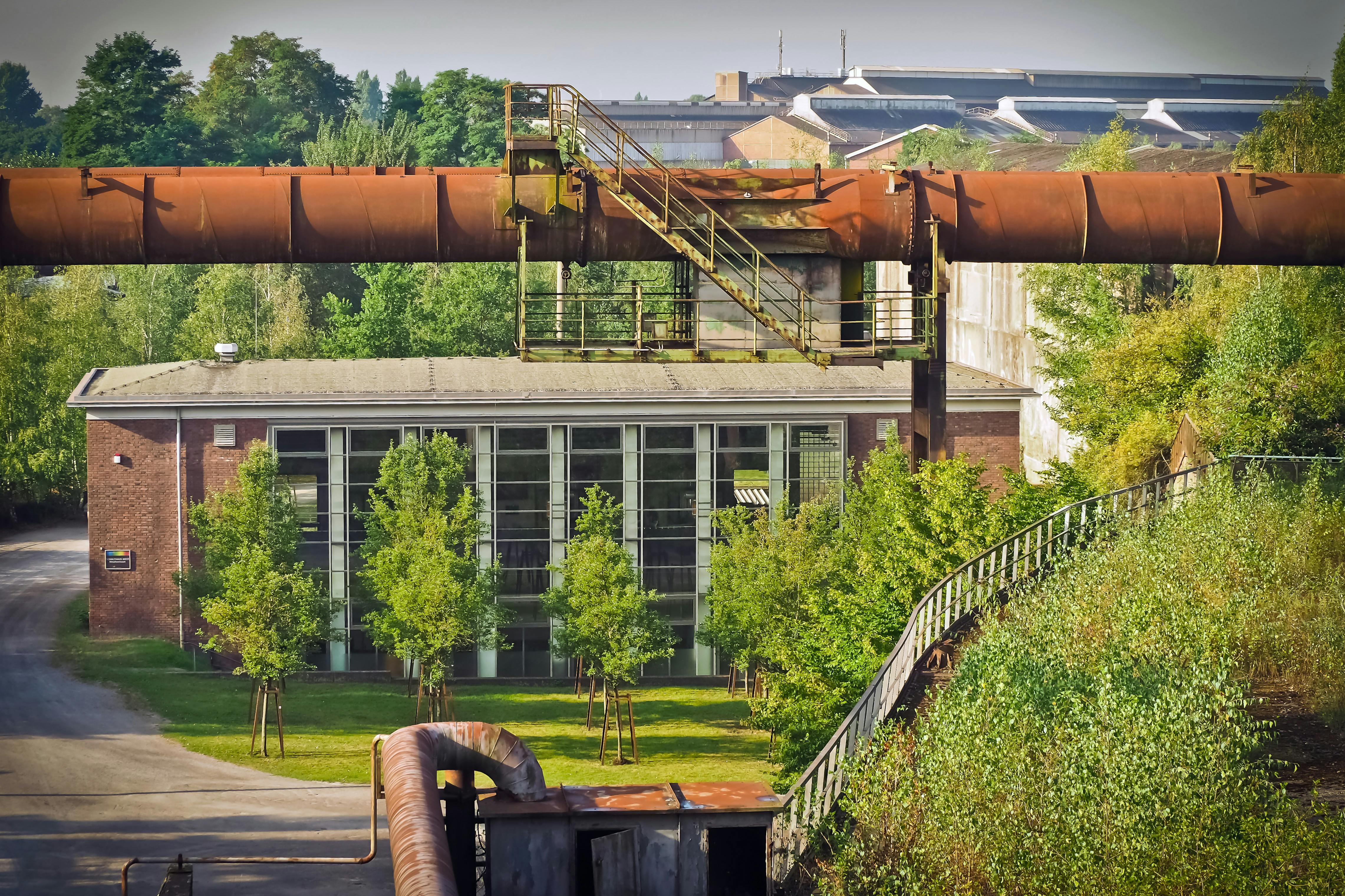 Ex struttura industriale riconvertita, con tubature arrugginite e verde che cresce intorno, simbolo di rigenerazione urbana e transizione sostenibile.