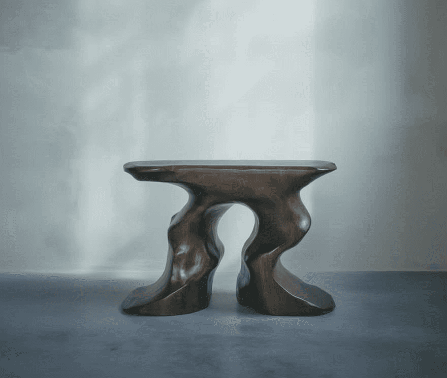 A modern, sculptural wooden table with an abstract, flowing design stands on a smooth, gray concrete floor against a softly lit, neutral-colored wall.