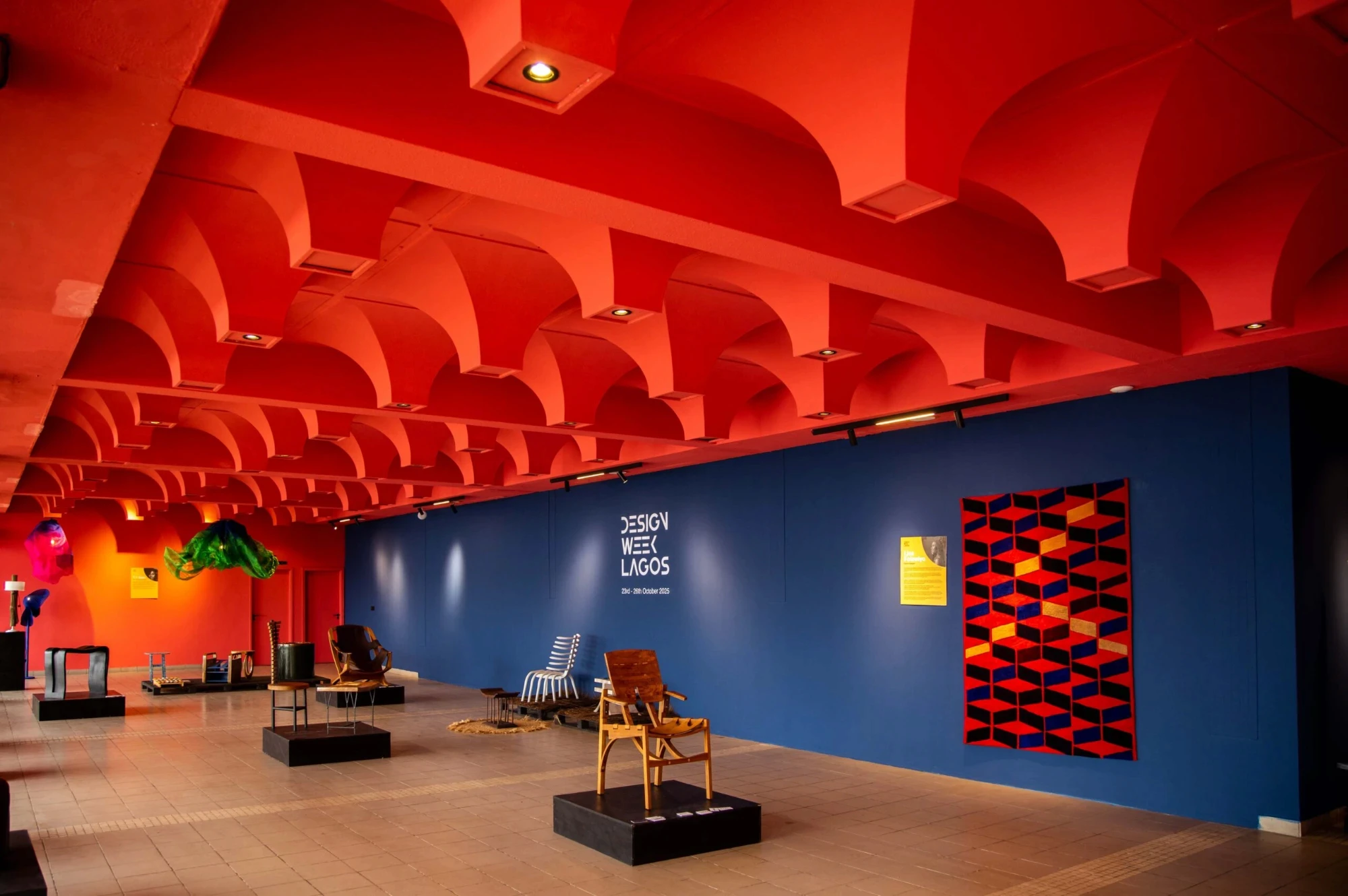 The Design Week Lagos 2025 exhibition: chairs, stools and a geometric textile on show under a red vaulted ceiling, beside a blue wall reading Design Week Lagos, 23rd-26th October 2025.