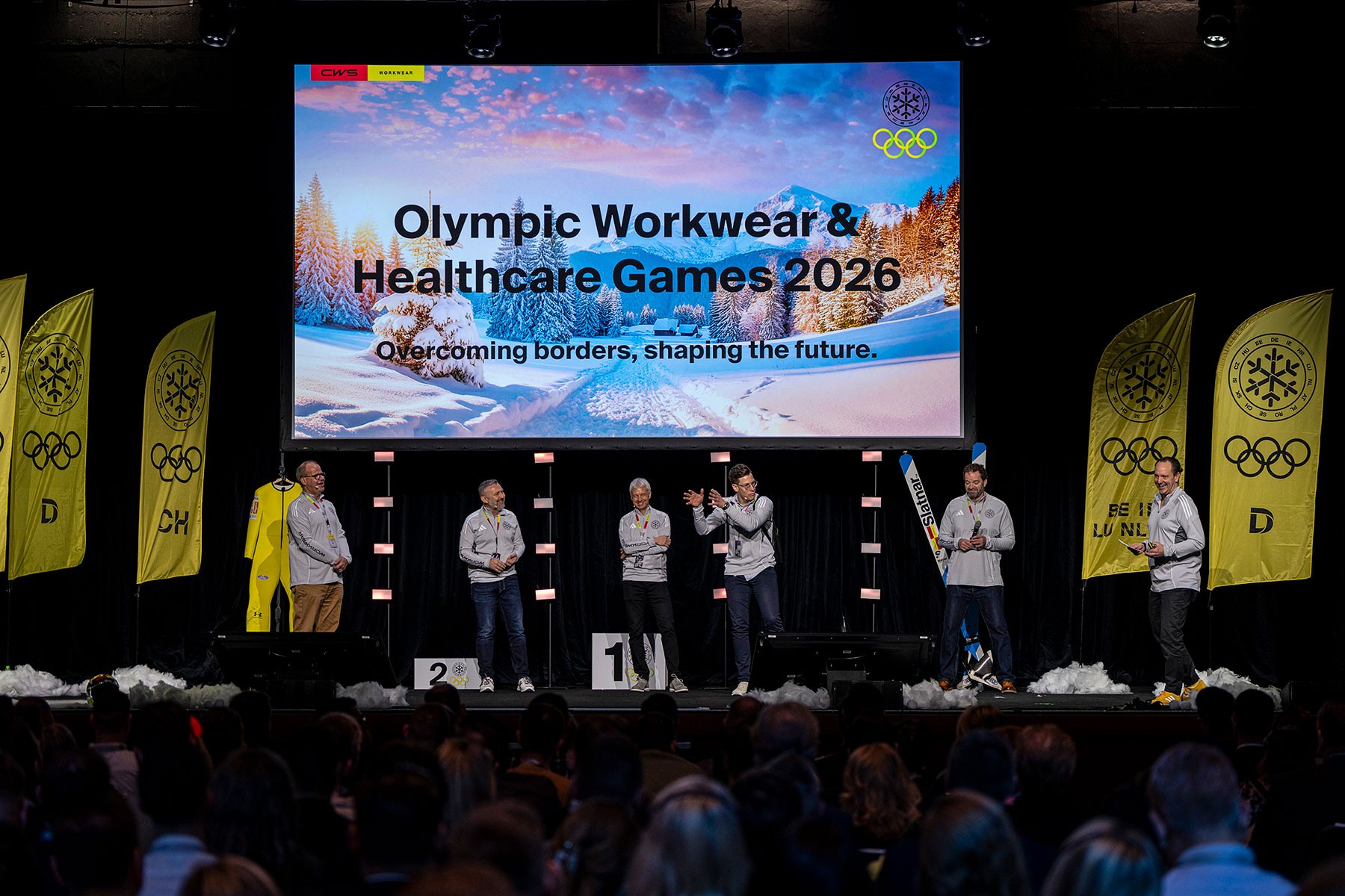 Group of speakers standing on the main stage during a corporate conference with a large audience present.