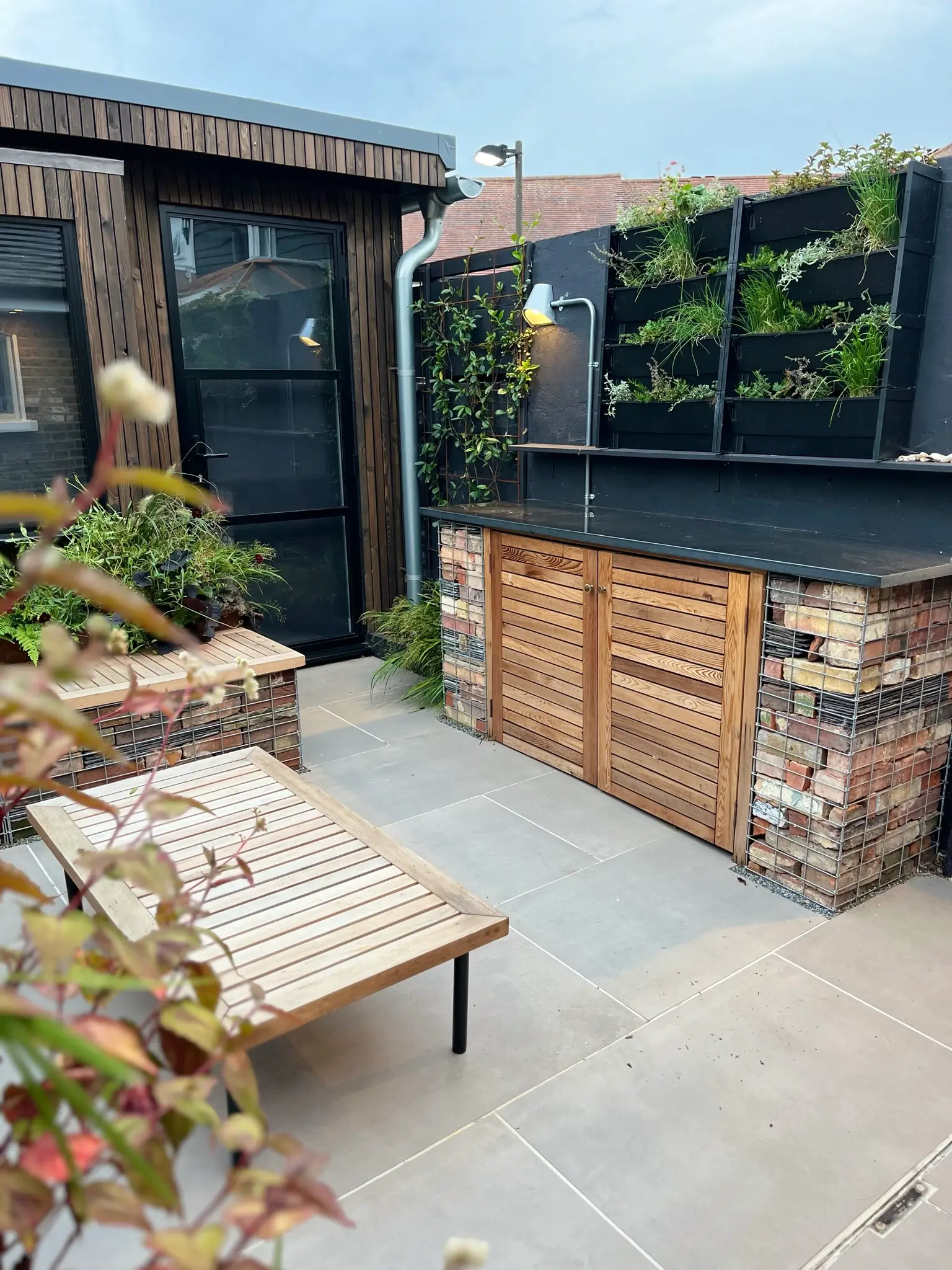 A cozy outdoor space featuring a wooden bench, a small table, and a vertical garden against a charming wall.