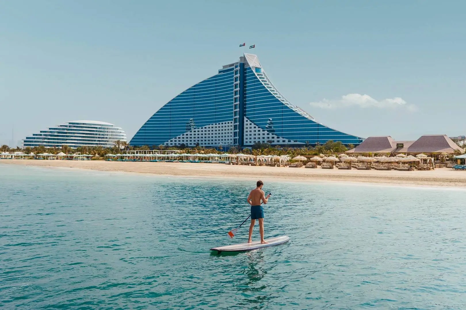 A man paddleboarding near the shoreline of Jumeirah, with the Jumeirah Beach Hotel and other buildings in the background.