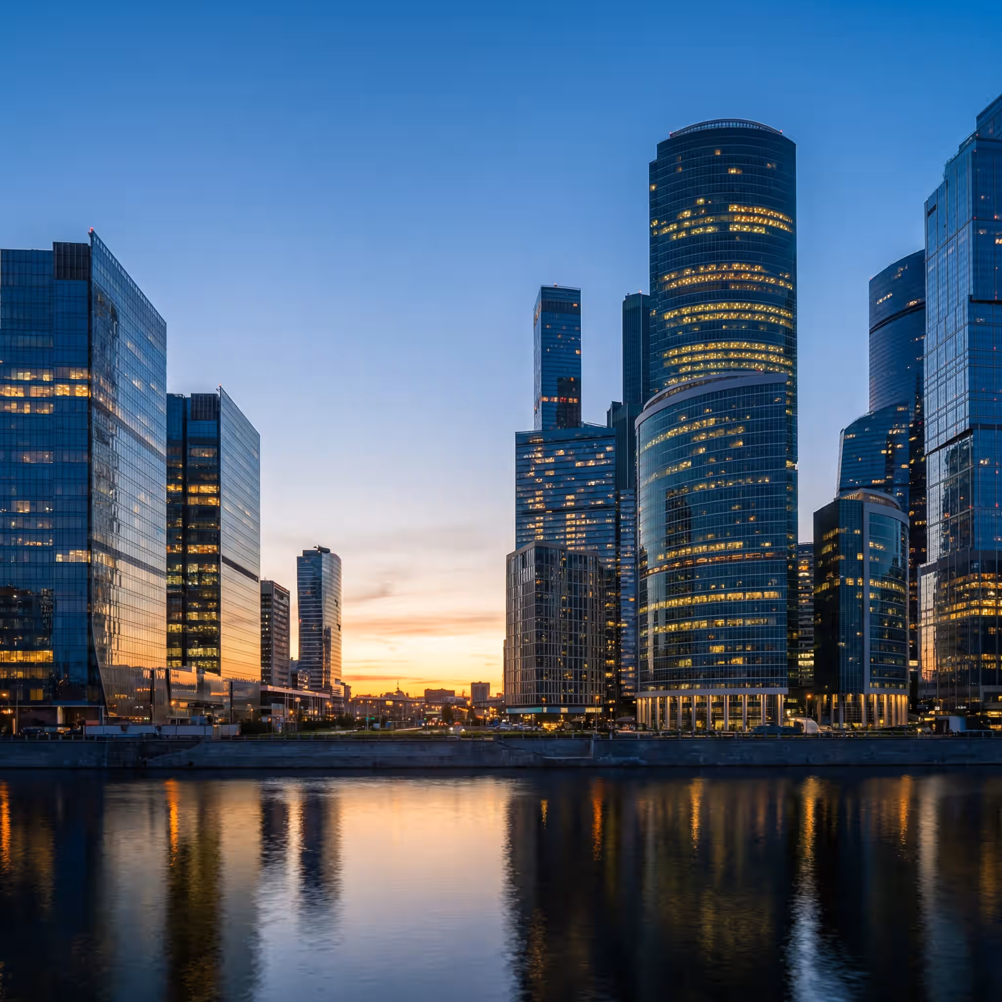 Cluster of illuminated glass skyscrapers reflected in a river at twilight under a deep blue sky.