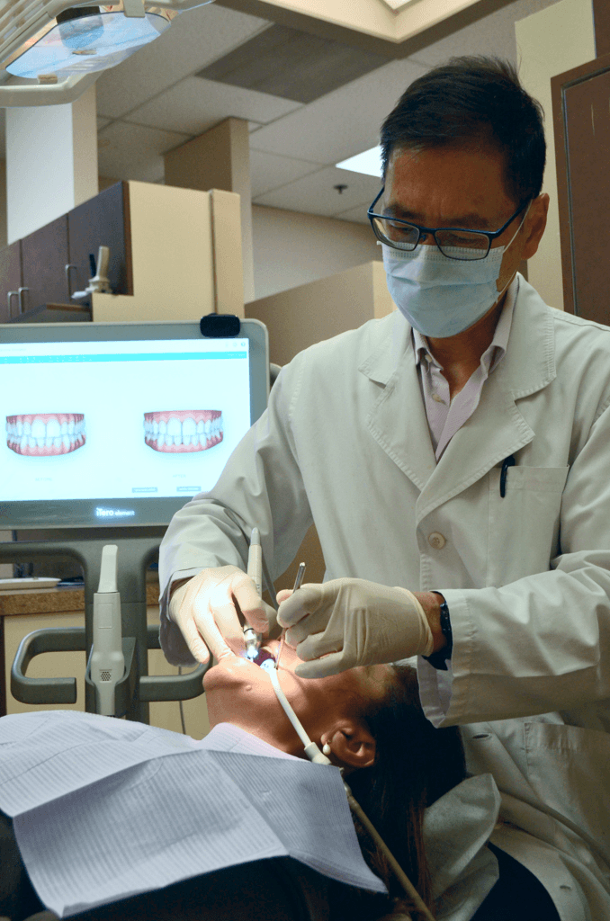 A dentist in a white coat and mask performing a dental examination, with a computer screen displaying dental images in the background.